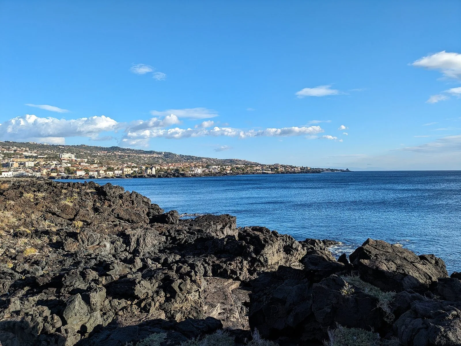 La scogliera di lava nera del lungomare di Catania verso Ognina, con il mare blu, un cielo azzurro con nuvole bianche e la costa costruita che sale verso le colline sullo sfondo