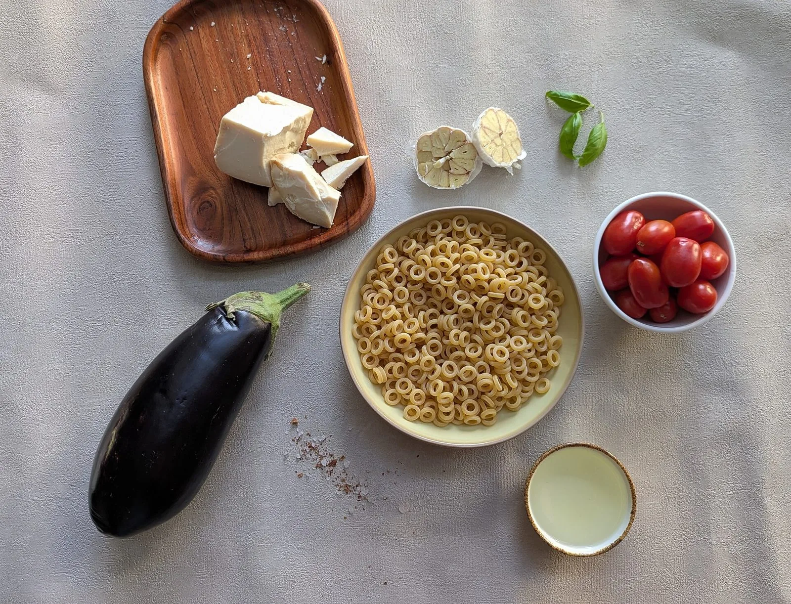 Gli ingredienti della pasta alla Norma disposti su un piano chiaro: una melanzana, una ciotola di anelletti crudi, scaglie di ricotta salata su un tagliere di legno, pomodorini, aglio, foglie di basilico e olio
