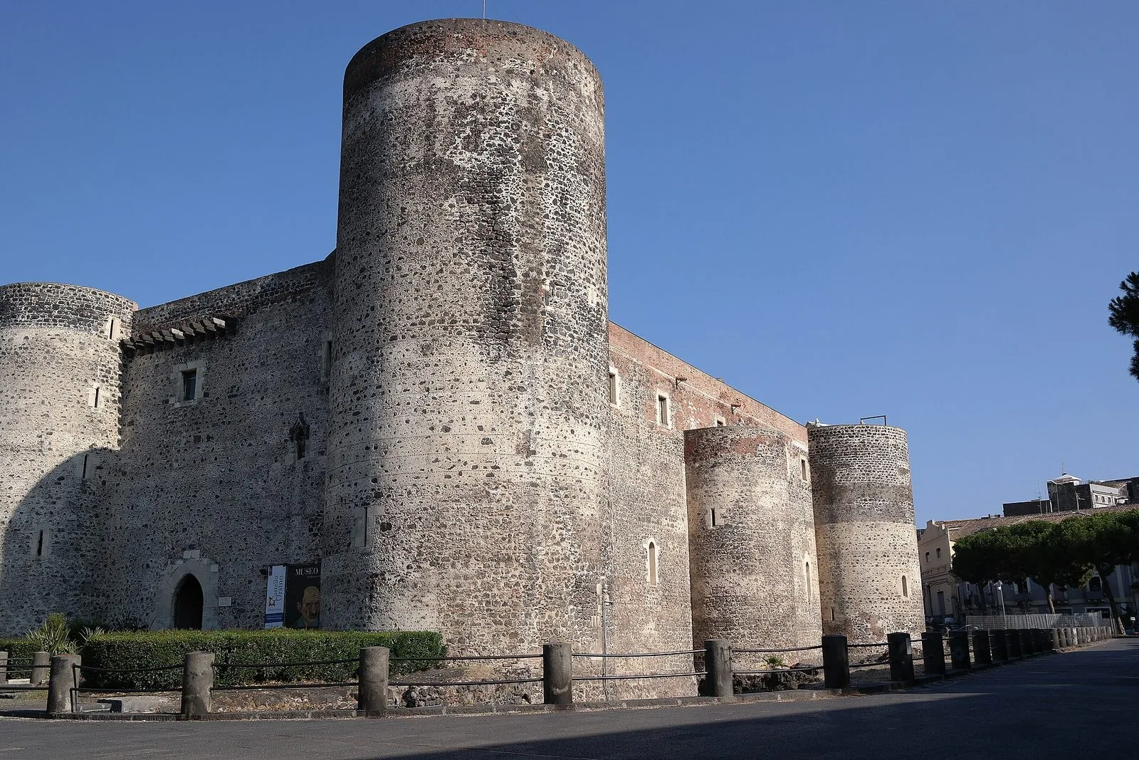 Il Castello Ursino di Catania con le sue torri cilindriche in pietra lavica grigia e nera sotto un cielo azzurro, con l'ingresso ad arco e una staccionata in primo piano