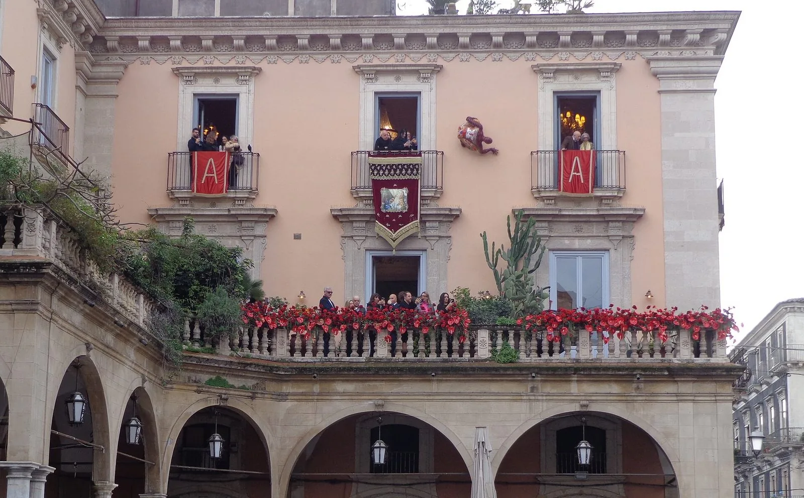 Facciata di un palazzo storico di Catania durante la festa di Sant'Agata, con drappi rossi con la lettera A ai balconi, uno stendardo con l'immagine della santa e una balconata decorata di fiori rossi sopra un porticato ad archi