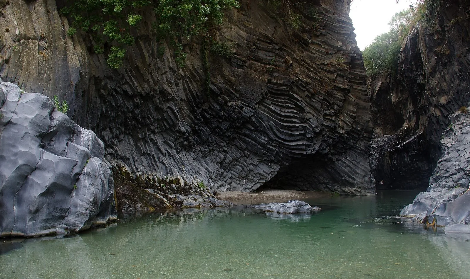 L'interno delle Gole dell'Alcantara: alte pareti di basalto grigio scuro con colonne prismatiche curve a ventaglio, una piccola grotta alla base e l'acqua limpida verde smeraldo del fiume in primo piano