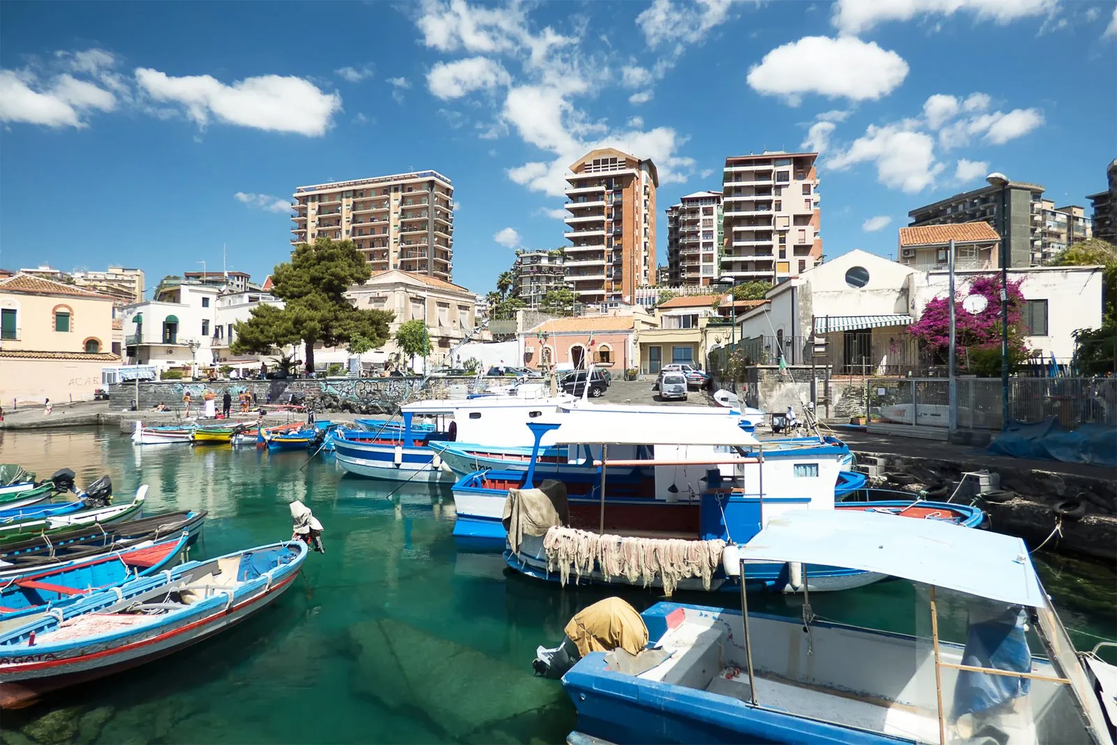 Il porticciolo di San Giovanni Li Cuti con barche da pesca bianche e azzurre ormeggiate nell'acqua verde, case basse e palazzi moderni sullo sfondo sotto un cielo con nuvole