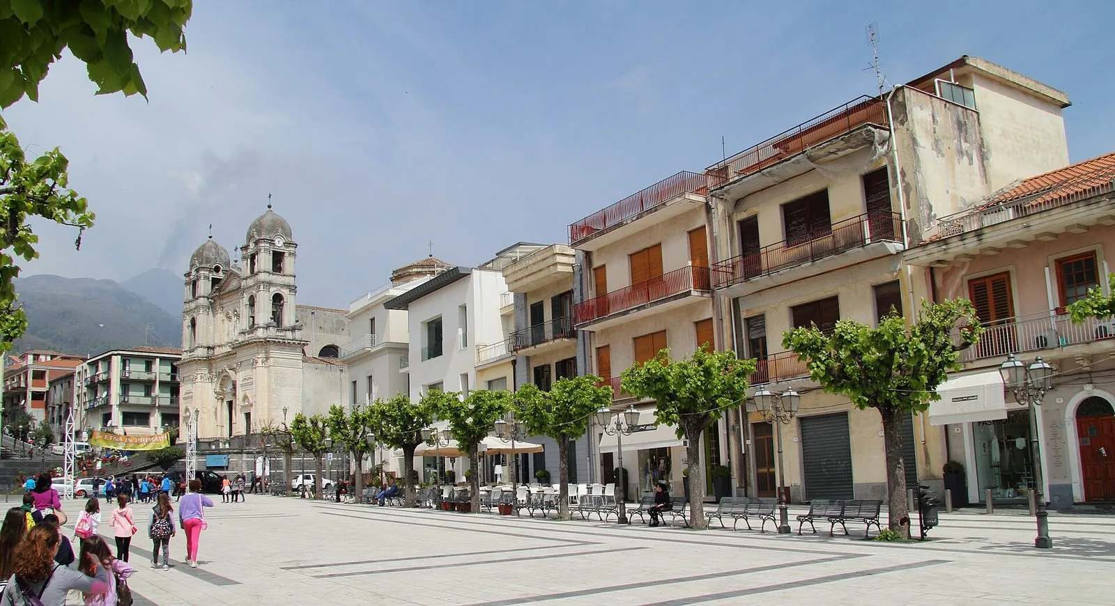 La piazza principale di Zafferana Etnea con la chiesa madre dalla facciata barocca a due campanili, una fila di palazzi con alberi potati e panchine, e le pendici dell'Etna con un pennacchio di fumo sullo sfondo