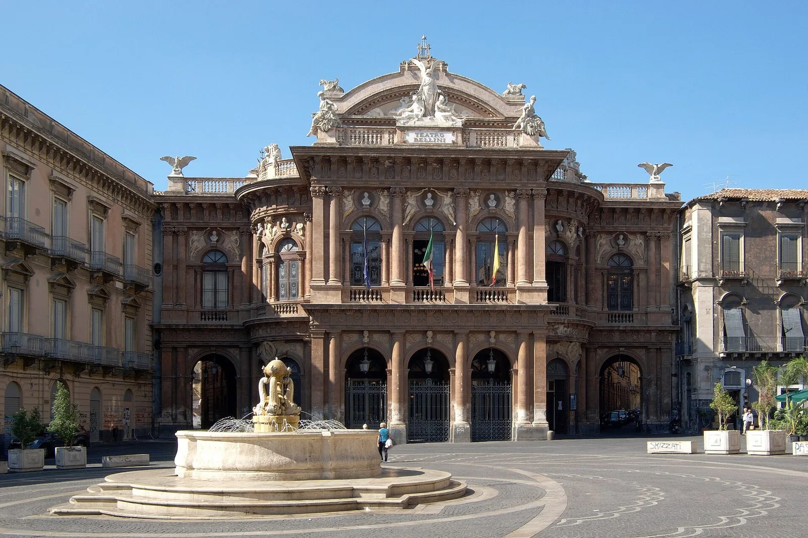 La facciata ottocentesca del Teatro Massimo Bellini di Catania sotto un cielo azzurro, con statue sul coronamento, colonne, balconi e una fontana in pietra al centro della piazza antistante